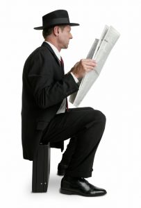 man in a hat reading a newspaper sitting on a briefcase